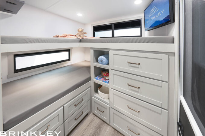 | Model Z 2810 Fifth Wheels 35 2026 Brinkley Model Z 2810 Fifth Wheel Rear Bunkhouse Interior featuring triple bunk beds, extensive white cabinetry with deep storage drawers and cubbies, a wall-mounted smart TV, and a private off-campside entry door.